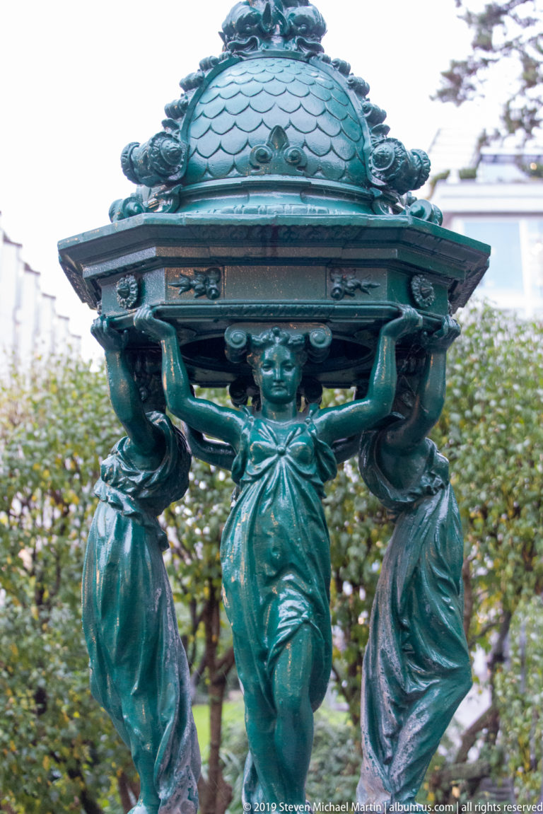 Three bronze-green female figures support an ornate domed basin or canopy in a Zurich public-space sculpture.