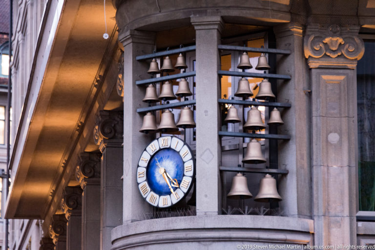 Blue Roman-numeral clock mounted between vertical rows of small bells on a stone Zurich façade.