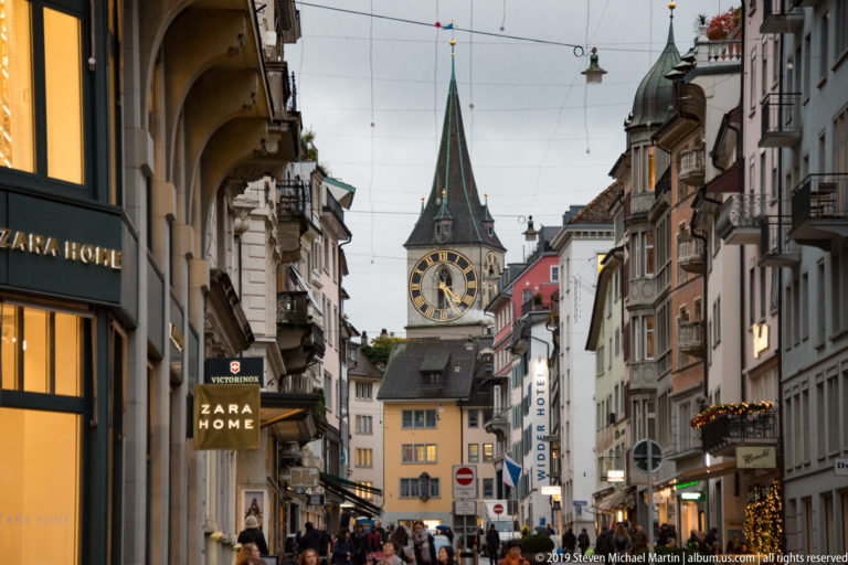 Busy Zurich Old Town street leading toward St. Peter’s tower and its large black-and-gold clock face.