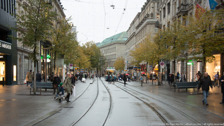 Bahnhofstrasse in Zurich with tram tracks and overhead wires, autumn trees, pedestrians, and illuminated shops.