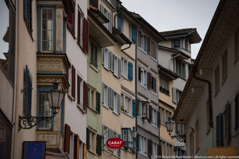 Narrow Zurich Old Town street lined with tall historic façades, multicolored shutters, lamps, signs, and a projecting oriel.