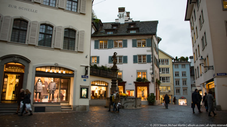 Stone figure fountain in a small Zurich pedestrian square surrounded by historic façades, lit shops, and pedestrians.