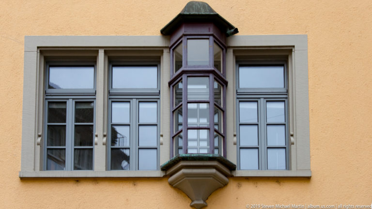 Narrow dark-framed polygonal oriel projects between tall windows on an ochre Zurich façade.