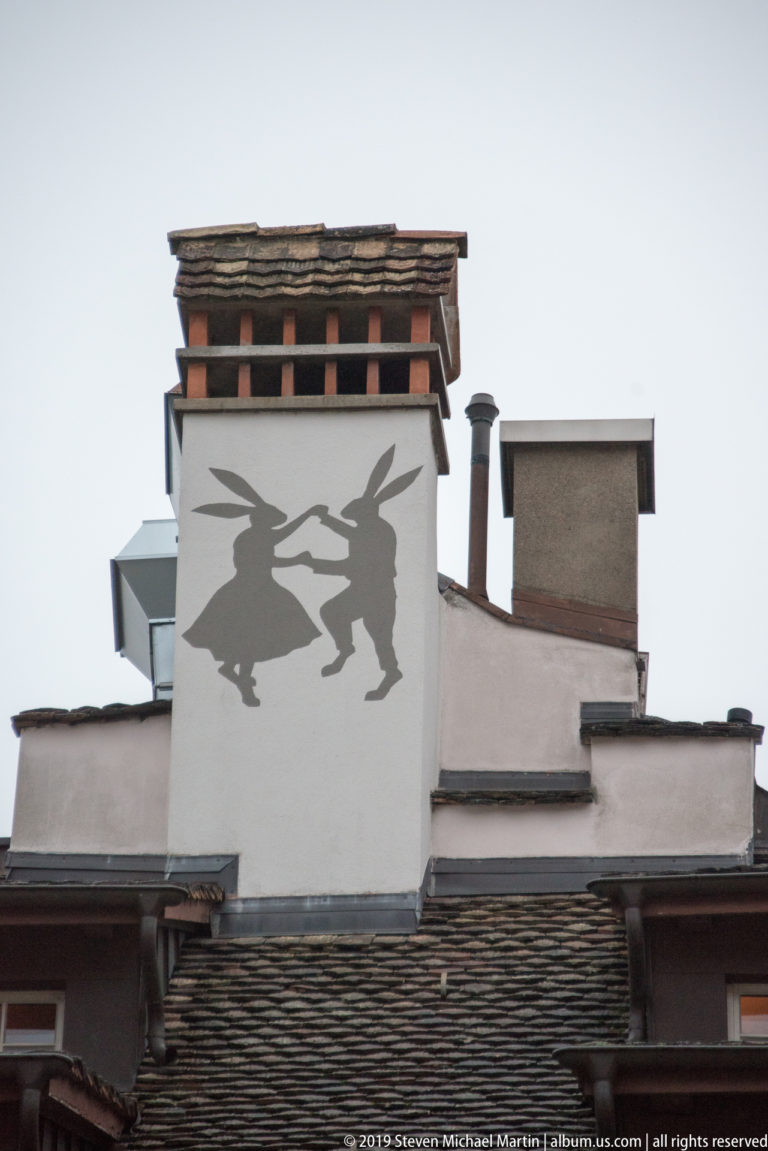 Two dark broom-riding figures painted on a white rooftop wall above tiled roofs and chimneys in Zurich.