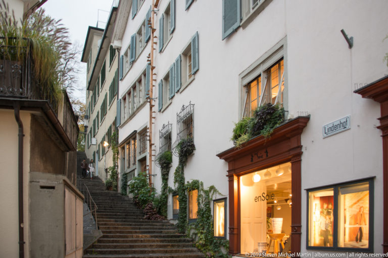 Stone steps climb a narrow lane marked Lindenhof between pale buildings with green shutters, vines, and shop windows.