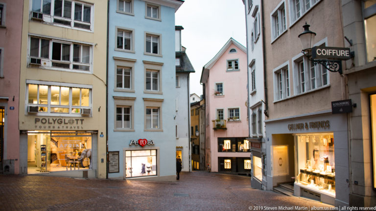 Small Zurich pedestrian space with pastel historic façades, illuminated shops, projecting signs, and a narrow passage beyond.