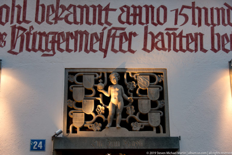 Red Gothic-script wall inscription above a dark figurative relief and blue number 24 plaque on a white façade.