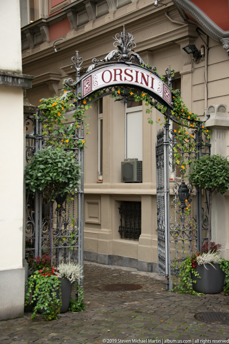 Ornate wrought-iron Orsini restaurant entrance with flowering vines, shrubs, and planters before a pale façade.