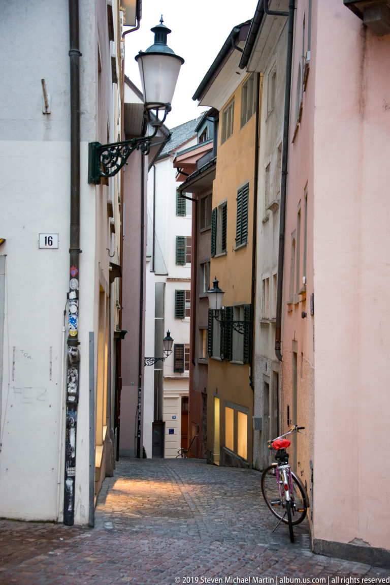 Cobblestone Zurich alley descending between pastel buildings, with shuttered windows, a streetlamp, and a parked bicycle.