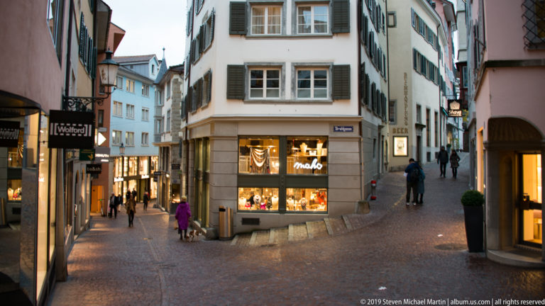 Narrow Zurich shopping lanes split around a corner jewelry store, with pedestrians and illuminated shop windows.