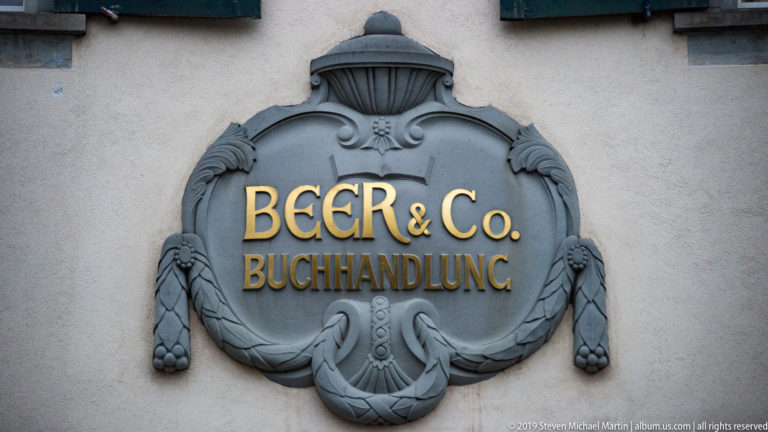 Gray ornamental wall plaque with gilded “BEER & Co. BUCHHANDLUNG” lettering on a pale façade.