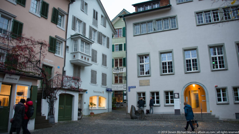 Pedestrians walk through a narrow Zurich Old Town lane lined with pale façades, shutters, bay windows, and shops.