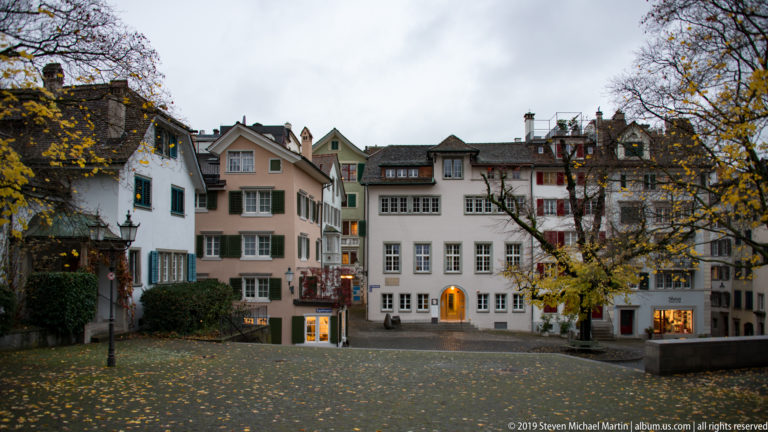 Leaf-strewn paved square bordered by narrow historic buildings, shops, and autumn trees in Zurich’s Old Town.