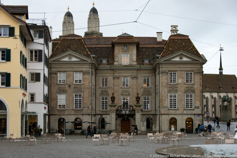 Pale symmetrical Zunfthaus zur Meisen façade with central balcony, tall windows, dormered roof, and outdoor seating in Münsterhof.