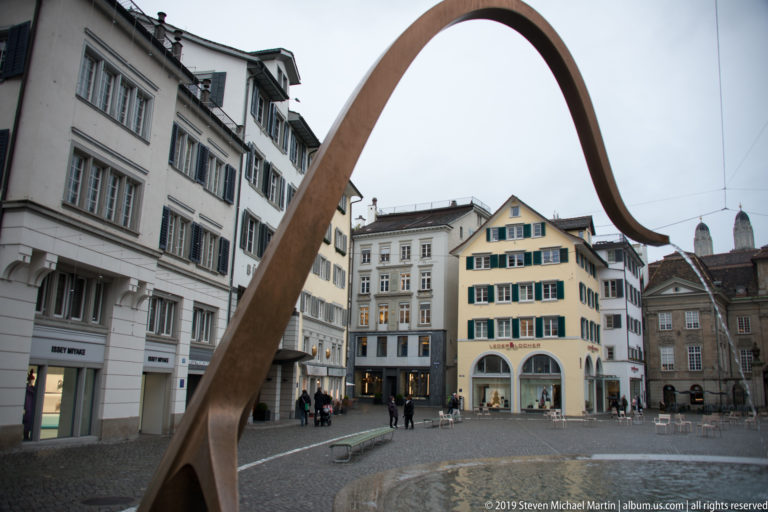 Large rust-colored curved steel sculpture arching over a paved Zurich square surrounded by historic buildings.
