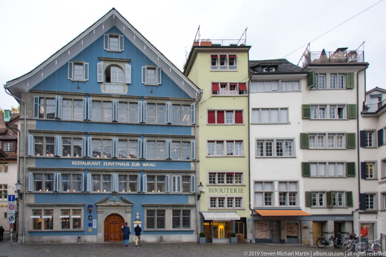 Blue Zunfthaus zur Waag façade with broad gable and rows of windows beside narrower historic houses in Zurich.