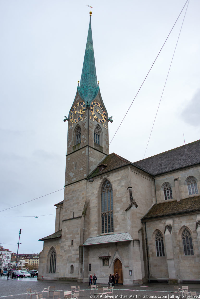 St. Peter’s Church with large clock tower, green spire, stone nave, Gothic windows, and tram wires overhead.