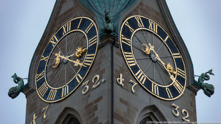 Two black-and-gold clock faces with Roman numerals on St. Peter’s stone tower beneath a green copper spire.