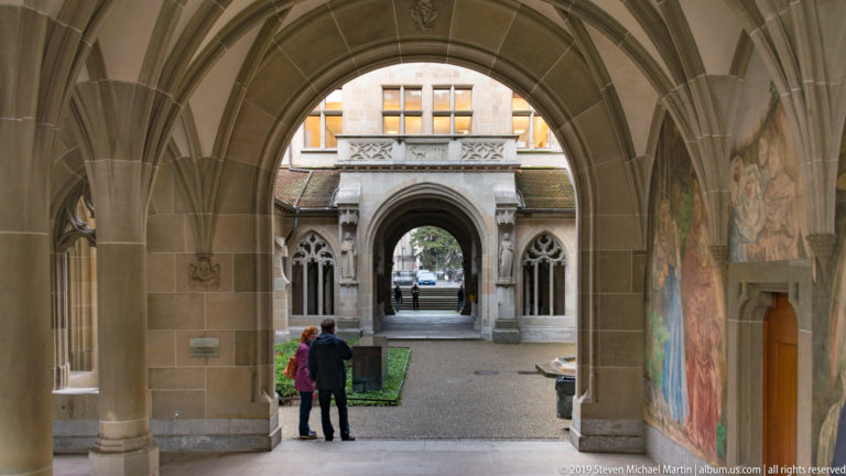 Two visitors beneath a stone cloister arch, with wall paintings, clustered columns, and successive courtyard openings beyond.