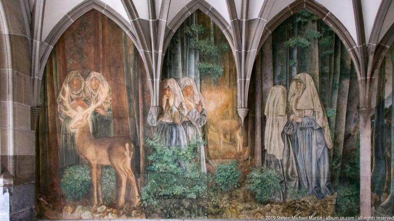 Cloister mural with robed women and a deer in a forest, divided by pointed stone arches.