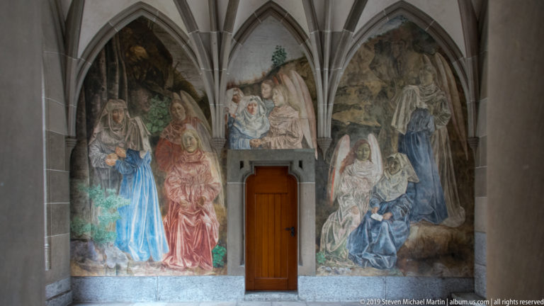 Narrative wall paintings of robed figures filling Gothic cloister bays around a central wooden door.