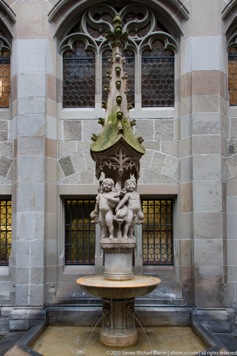 Sculptural courtyard fountain with clustered figures, steep canopy, and circular basin before Gothic traceried windows.
