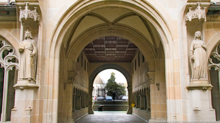 Pointed Gothic cloister entrance with standing stone figures on both sides and a passage opening toward a courtyard.