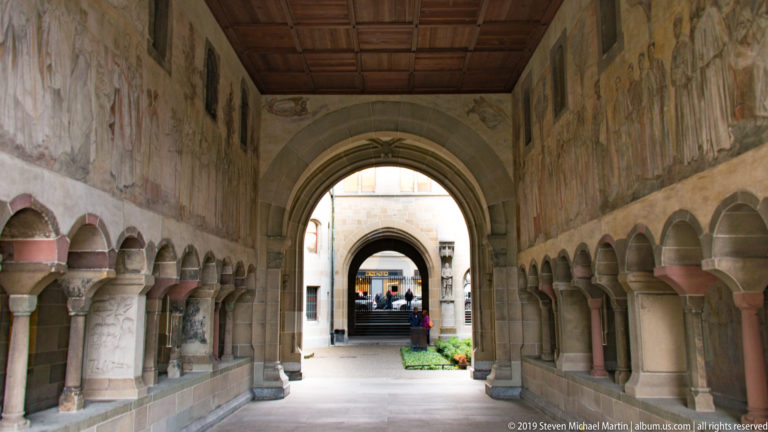 Long Zurich cloister passage with paired stone columns, rounded arches, wall paintings, and a coffered wooden ceiling.
