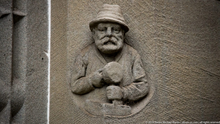 Small stone relief of a bearded man in a broad cap holding an object at chest height.