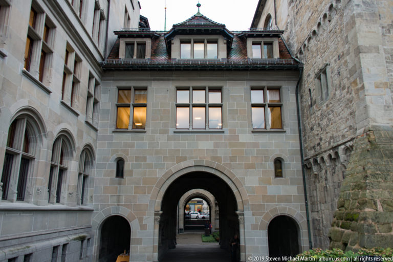 Large stone archway leading through a covered passage toward a courtyard, with smaller side arches in a Zurich cloister.