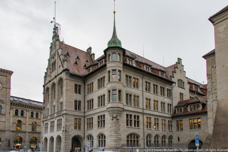 Ornate stone corner building with arched openings, steep roof, carved masonry, and small green copper turret in Zurich.