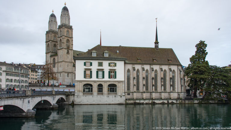 Wasserkirche beside the Limmat with tall Gothic windows, bridge arches, and a Grossmünster tower rising behind.