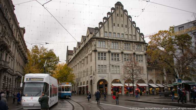 Route 7 tram approaching on curved tracks through a Zurich square with pedestrians, autumn trees, and a stepped-gable building.
