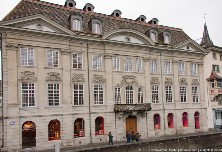 Symmetrical pale-stone Zurich façade with tall windows, carved lintels, central balcony, and elaborate roofline.
