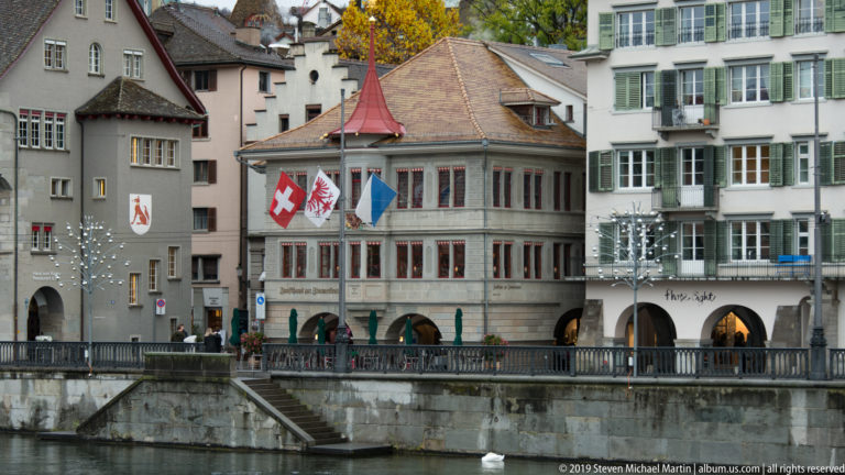 Zunfthaus zur Saffran on the Limmat with flags, arcaded ground floor, red-trimmed windows, and steep roof.