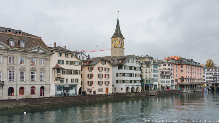 St. Peter’s clock tower and green spire rising above historic buildings along the Limmat in central Zurich.