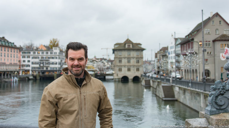 Man in tan jacket beside the Limmat with Zürich Rathaus and historic riverfront buildings behind him.