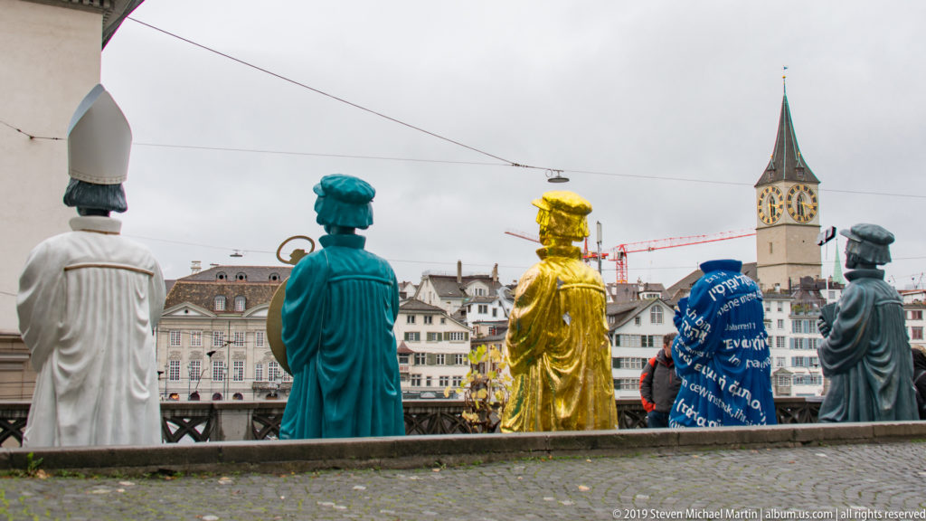 Brightly colored sculptural figures seen from behind with St. Peter’s clock tower rising over Zurich’s Old Town beyond.
