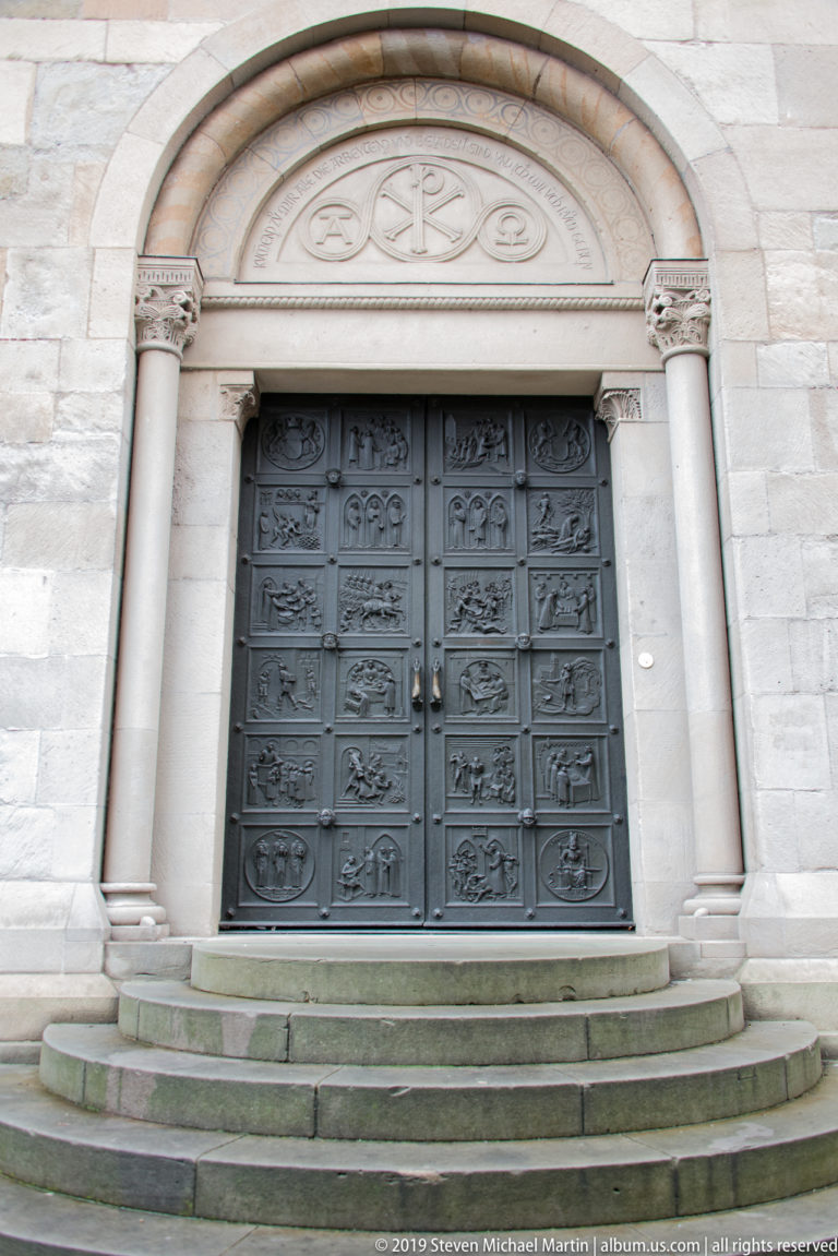 Dark relief-covered double doors within a pale stone church portal, flanked by columns and approached by shallow steps.