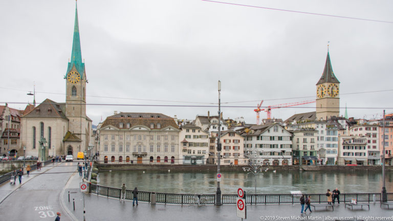 Fraumünster spire and St. Peter’s clock tower rising above historic buildings across the Limmat in Zurich.