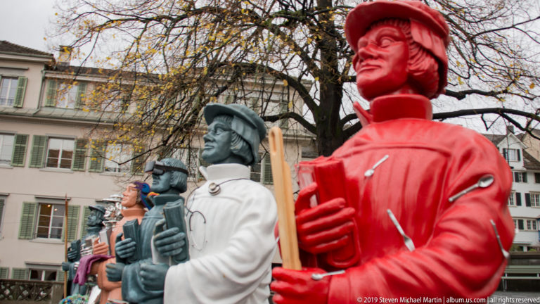 Row of oversized brightly colored figures in historical-style clothing beneath autumn trees in Zurich.