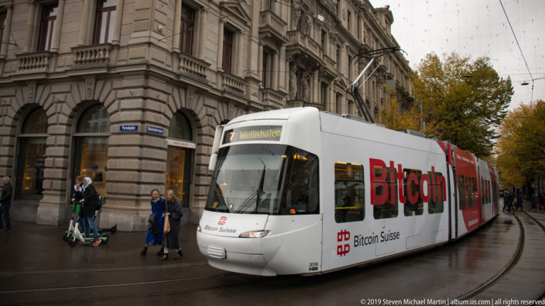 White articulated Zurich tram on route 7 to Wollishofen, wrapped in Bitcoin Suisse advertising, curving past historic buildings.