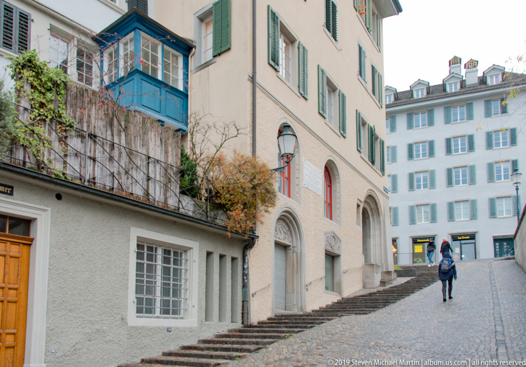 Steep cobblestone Zurich lane with side steps, green shutters, arched openings, climbing plants, and a lone pedestrian.