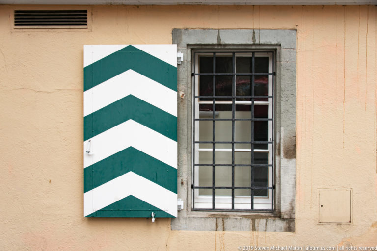 Open green-and-white chevron shutter beside a narrow barred window in a gray stone frame on a peach wall.