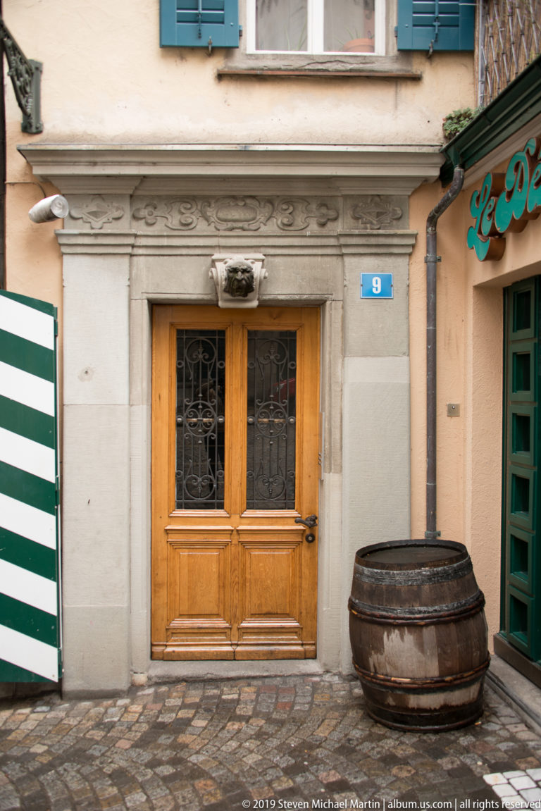 Wooden double door in a carved stone surround with small animal-head ornament, barrel, cobblestones, and striped shutters.