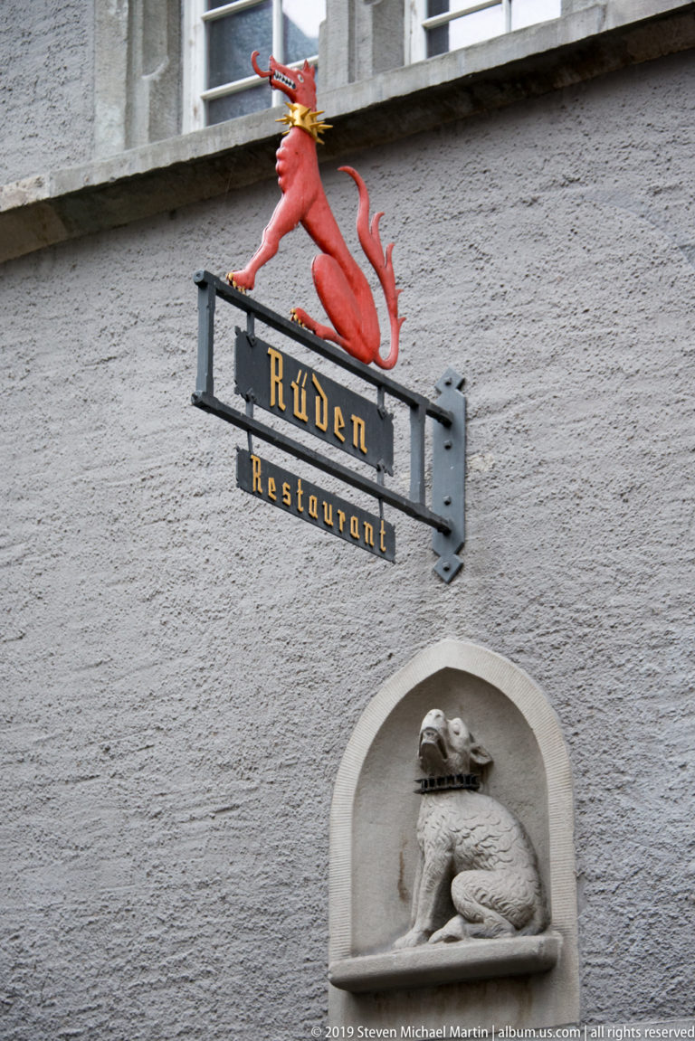 Red hound above a black Rüden Restaurant sign, with a carved curled dog in a wall niche below.