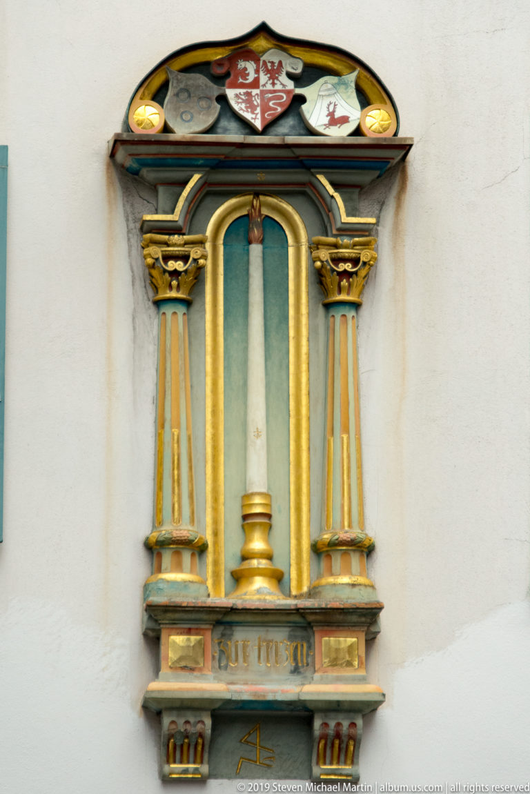 Small gilded wall niche with paired columns, turquoise recess, heraldic crest, and inscription on a Zurich façade.