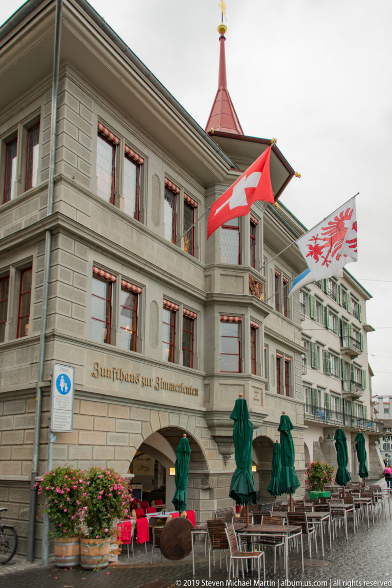 Zunfthaus zur Zimmerleuten with red-trimmed windows, arcades, Swiss and heraldic flags, and outdoor tables.