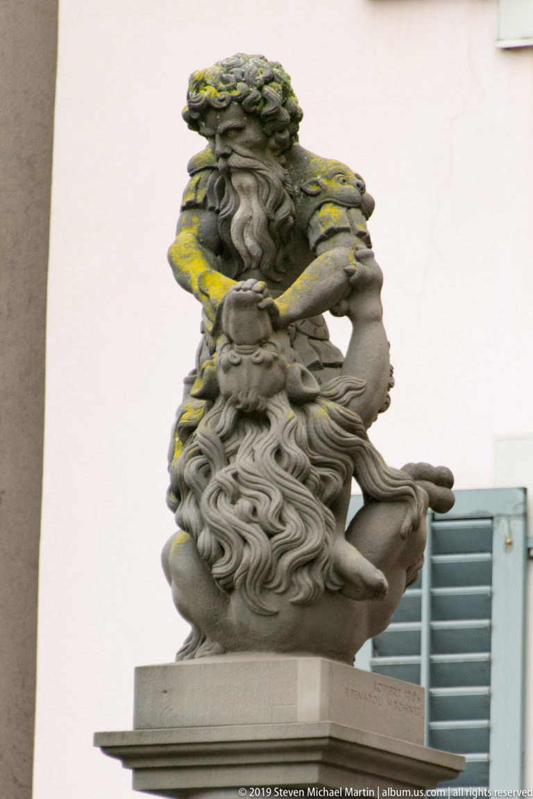 Weathered stone sculpture of a bearded seated figure holding a smaller figure, with moss across the hair and shoulders.