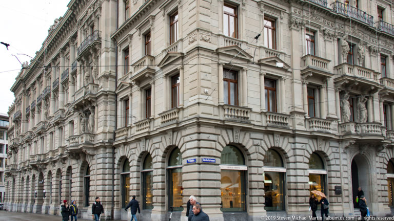 Ornate pale-stone corner building with arched shops, balconies, carved window surrounds, and pedestrians on Bahnhofstrasse in Zurich.